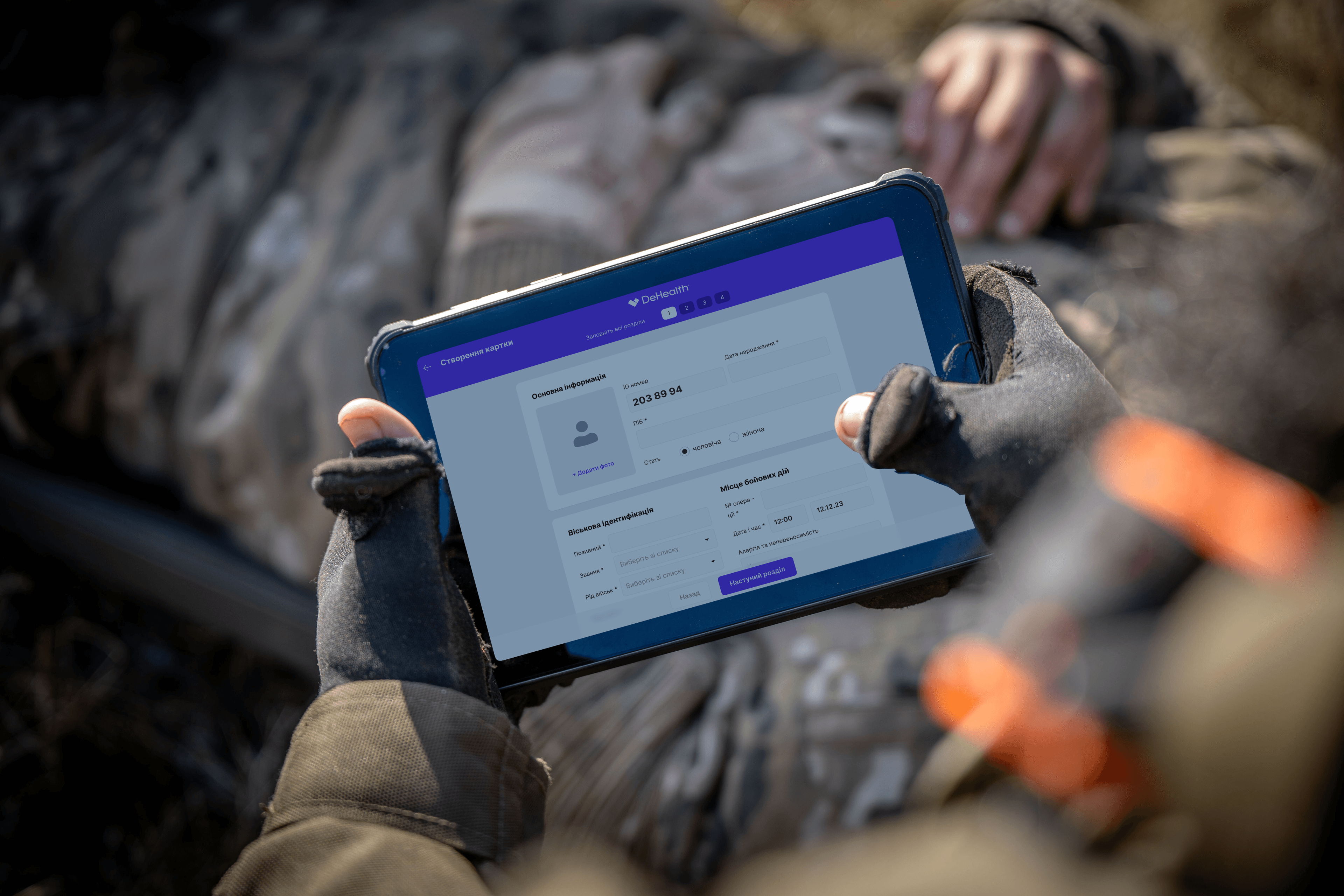 Combat medic creating a casualty record on the AHS-UA1 tablet — DeHealth interface visible, wounded soldier in the background